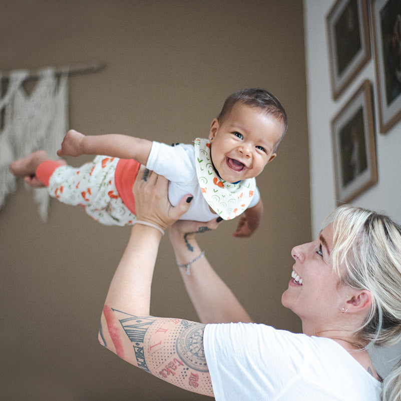 Mutter hebt lachendes Baby hoch in die Luft und lächelt es an.