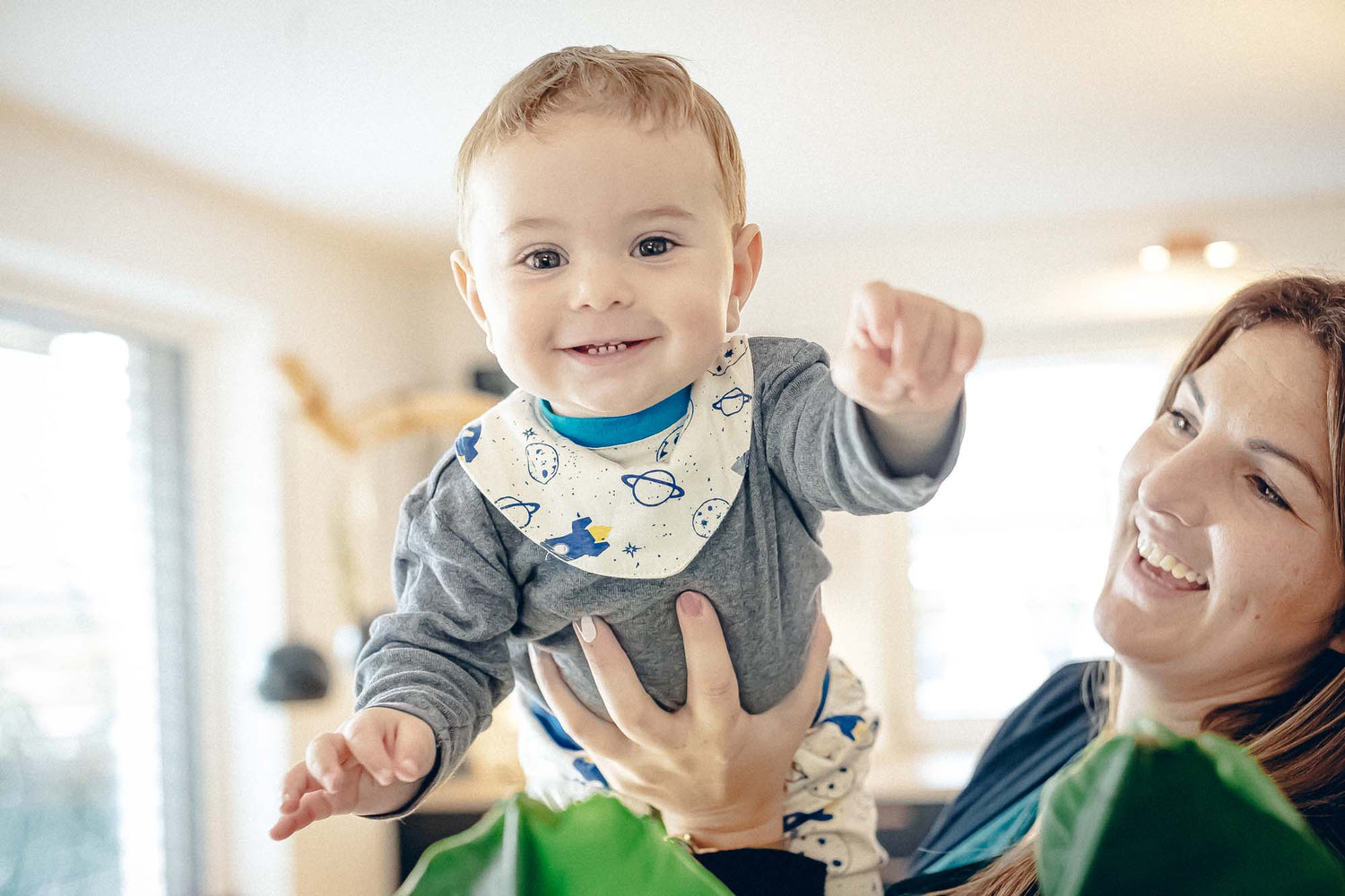 Mutter hält Baby hoch. Baby trägt Windelbär Spucklätzchen und lächelt.