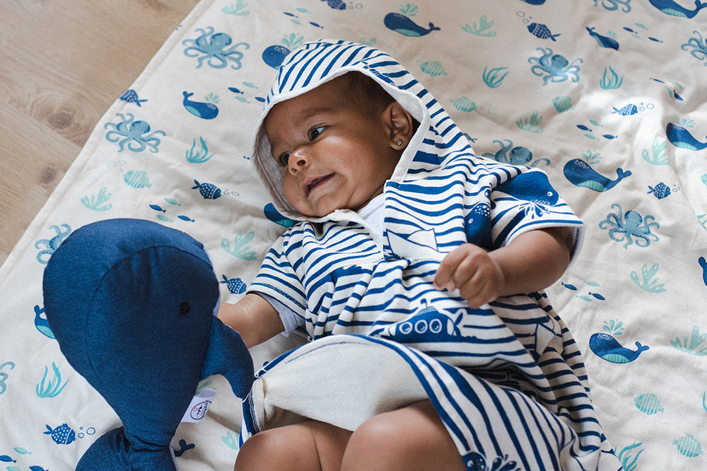 Baby liegt in Baderobe auf Babydecke von Windelbär Design Fische & Boote blau und spielt mit Kuscheltier.