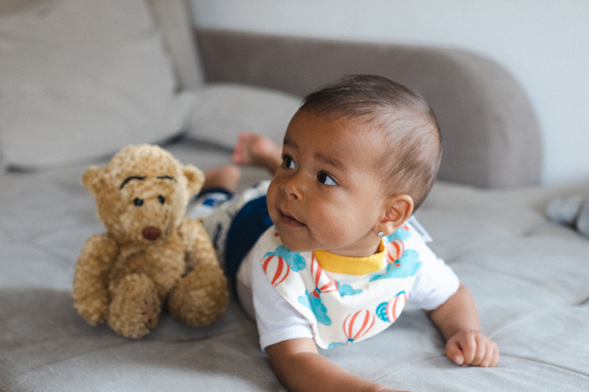 Baby mit Windelbär Spucklätzchen liegt neben einem Kuscheltier auf dem Bauch und stützt sich auf den Armen ab.